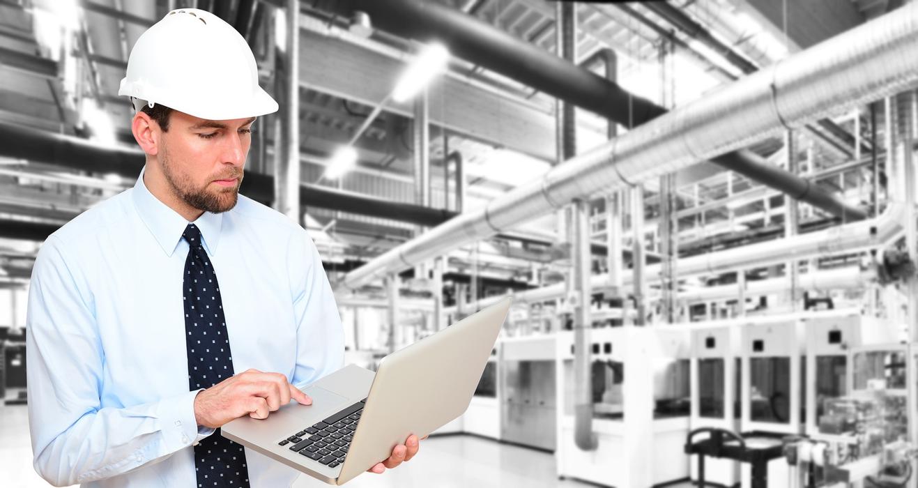 Mitarbeiter steht in einer Produktionshalle mit einem Laptop in der Hand
