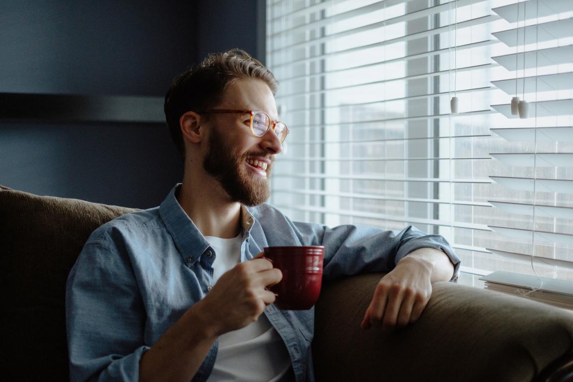 Mann schaut lächelnd mit roter Kaffeetasse in der Hand aus Fenster